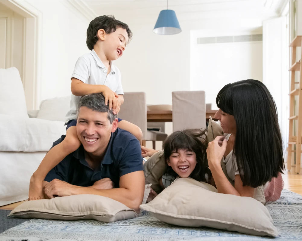 Padre, madre e hijos pequeños jugando sonrientes en casa, representando seguridad y estabilidad económica con un seguro de vida.